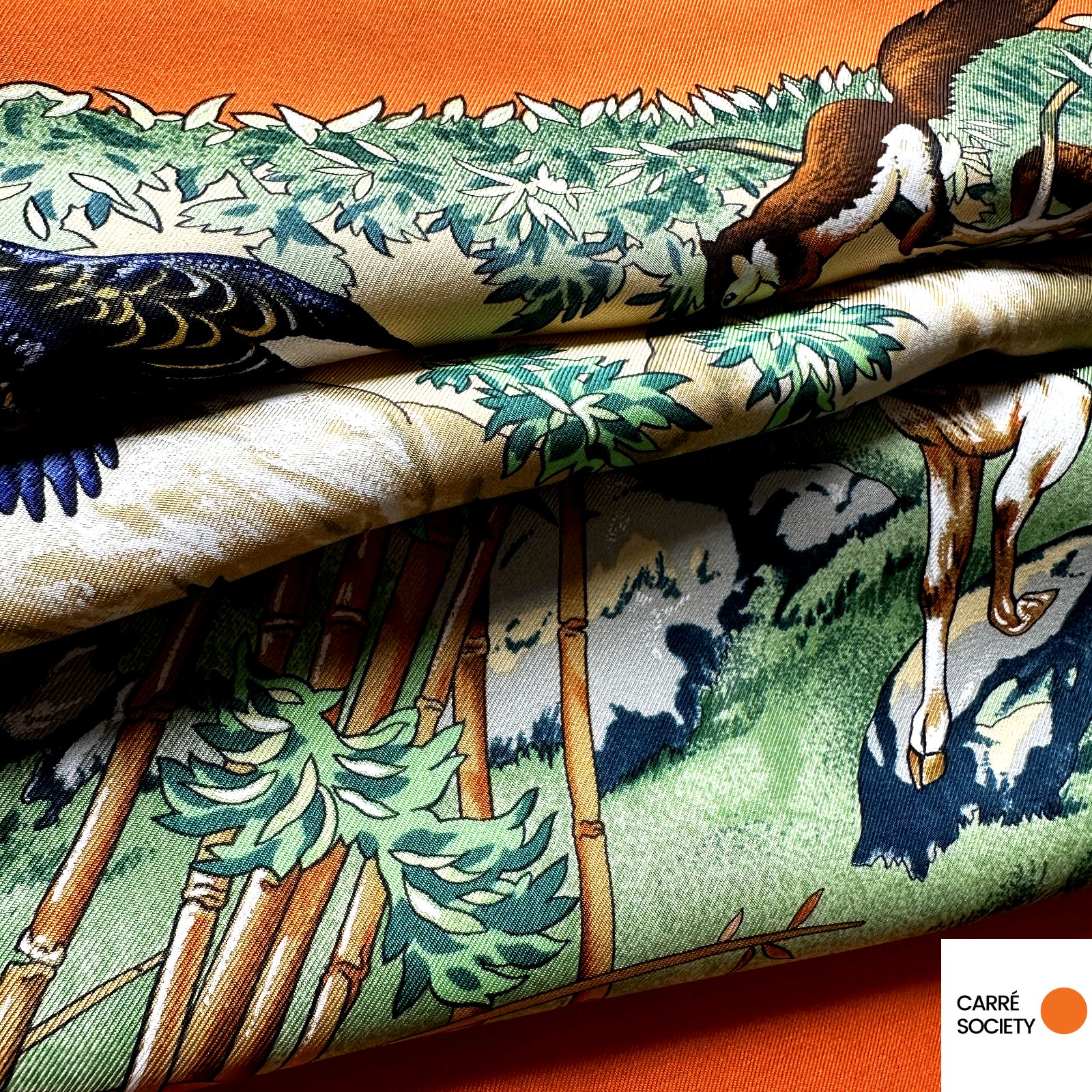 foulard replié, texture soyeuse, bordure orange vif contrastant avec les tons verts.